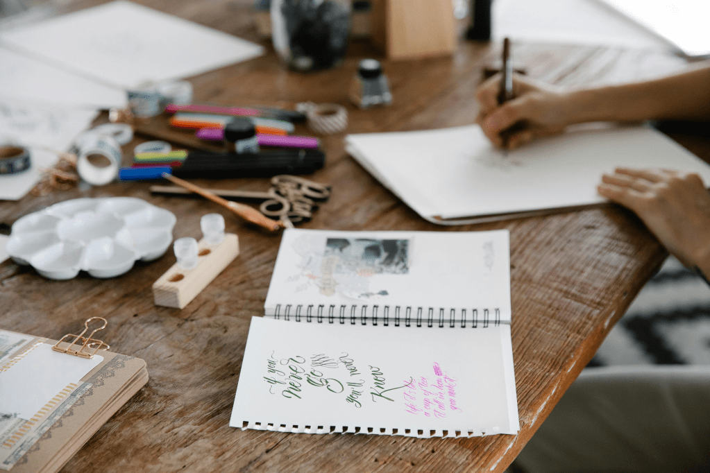 Tisch, Blätter und Stifte. Eine Hand, die auf ein Papier schreibt. 